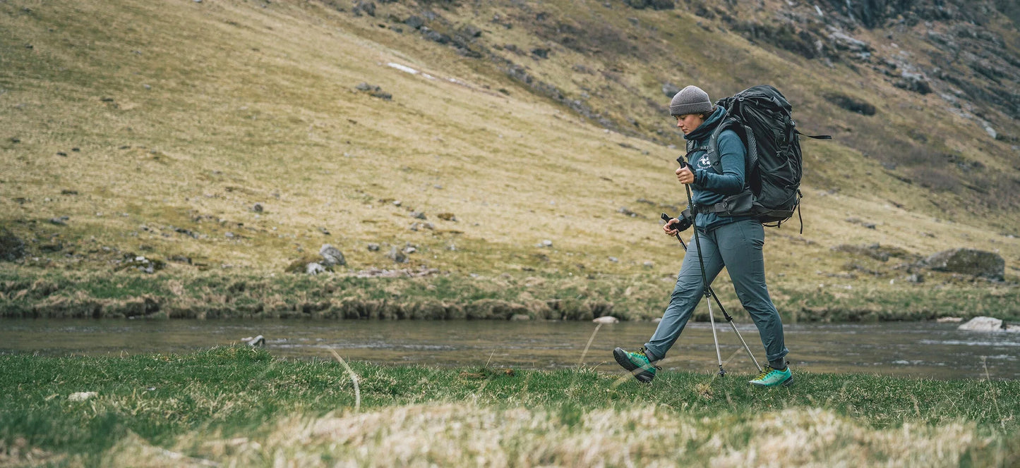 Hiker with backpack and trekking poles walking near river in mountainous terrain, outdoor adventure gear