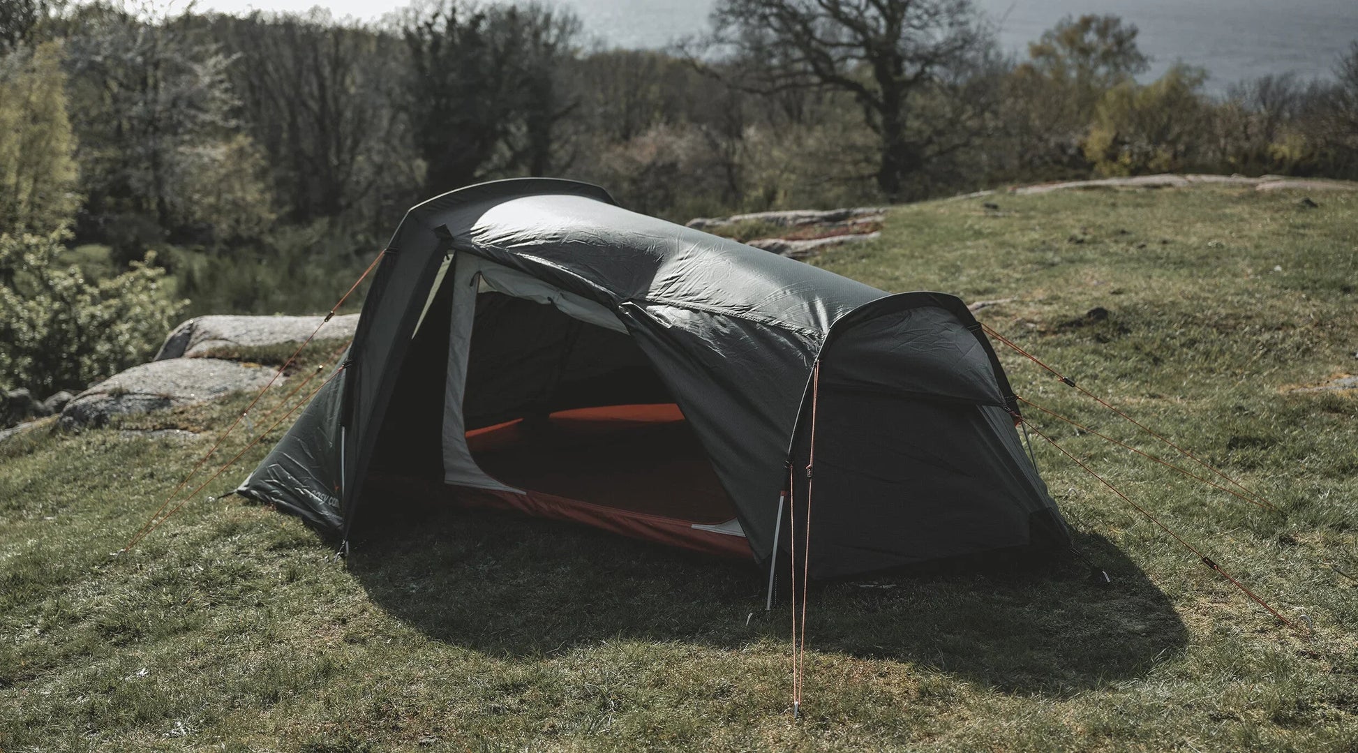 Gray camping tent set up on grassy hillside with trees and lake view, ideal for outdoor adventures.