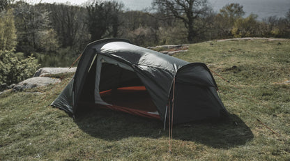 Gray camping tent set up on grassy hillside with trees and lake view, ideal for outdoor adventures.
