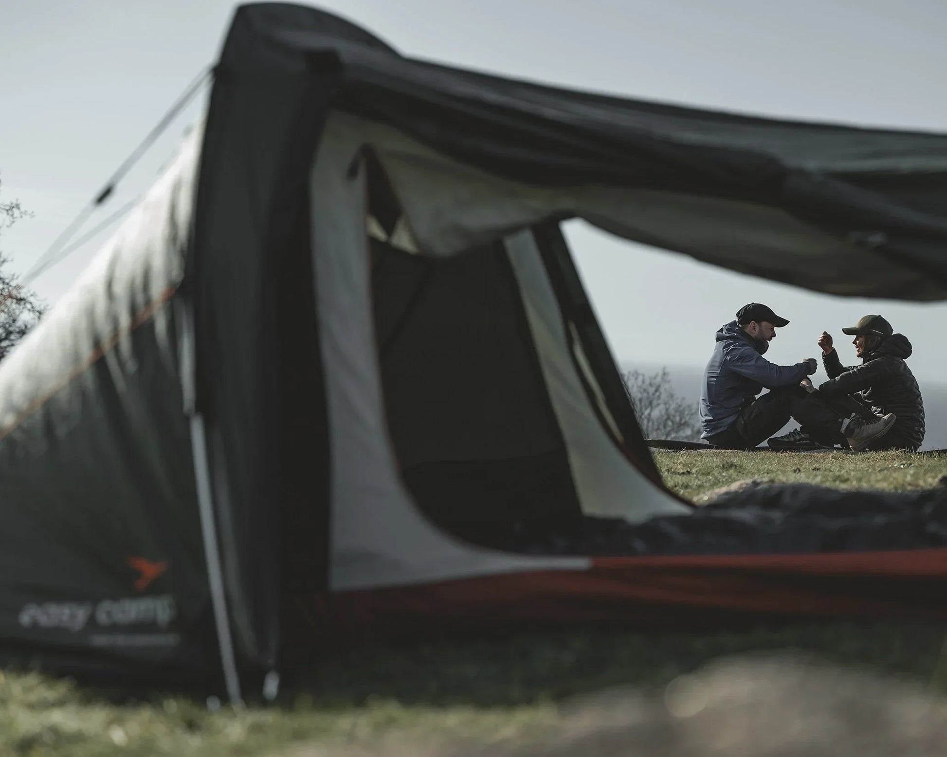 Couple relaxing outside a camping tent in outdoor gear, enjoying nature on a grassy hill.