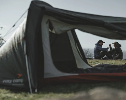 Couple relaxing outside a camping tent in outdoor gear, enjoying nature on a grassy hill.