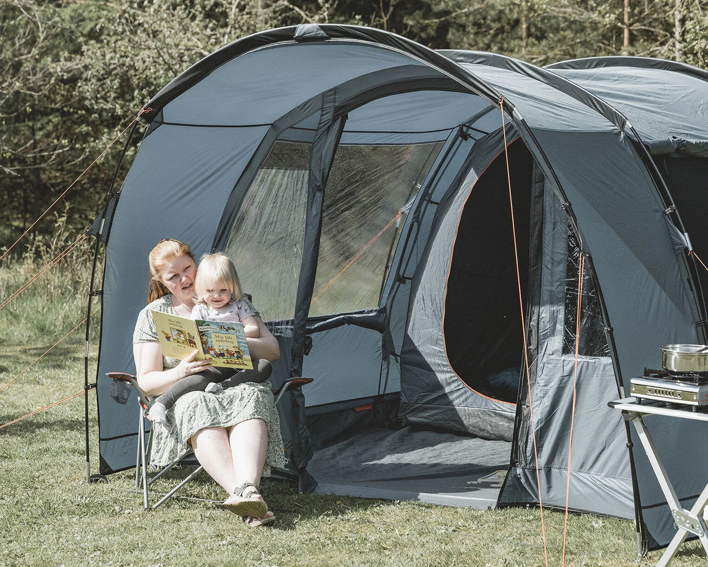 Family camping with large grey tent, portable stove, and folding chairs in grassy outdoor setting