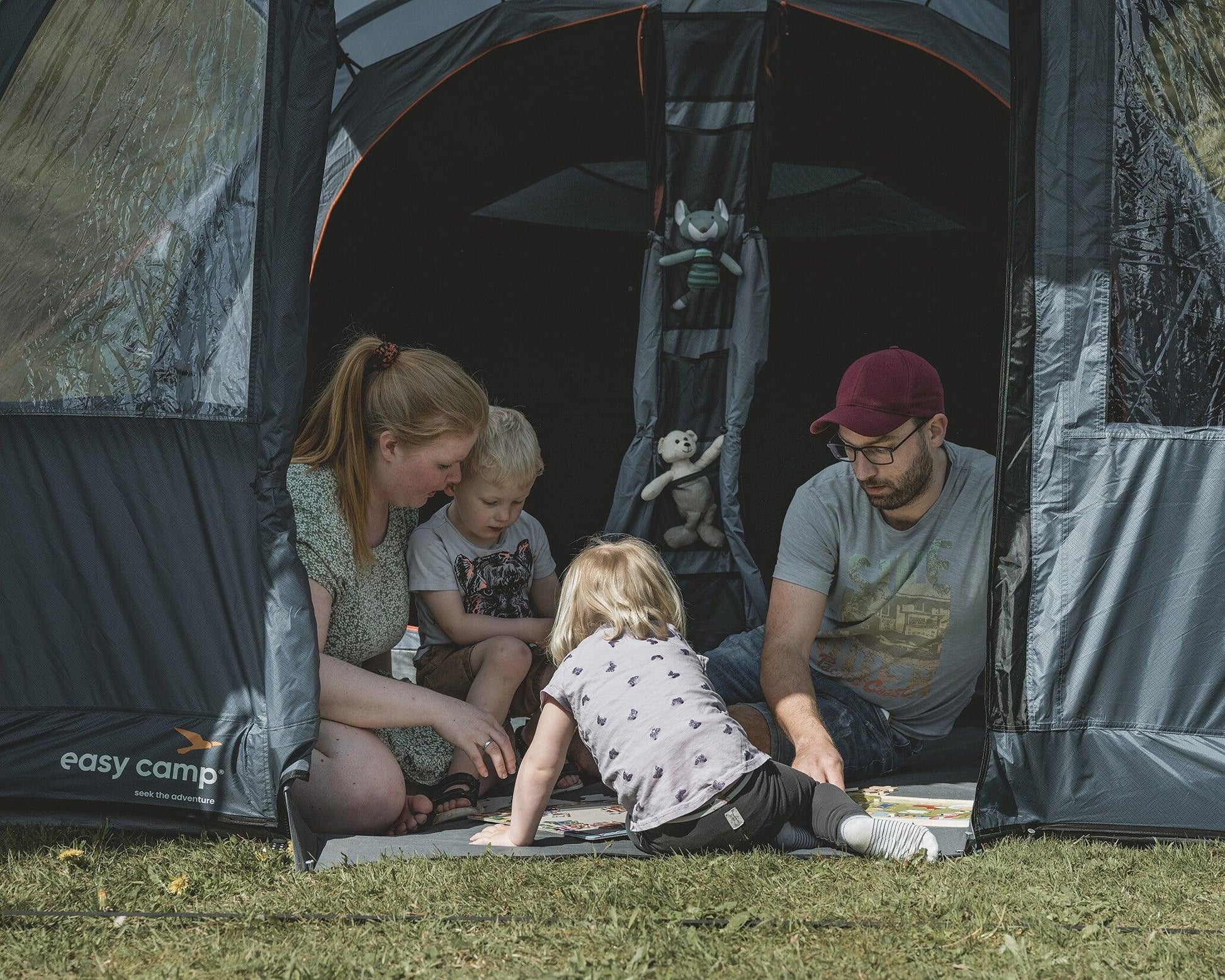 Family enjoying camping inside a tent with kids playing games and plush toys hanging inside.
