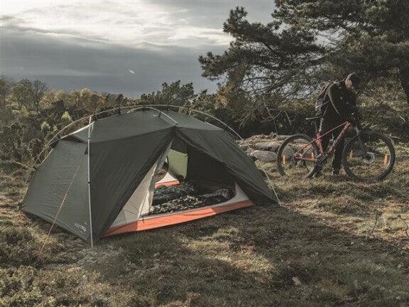 Green camping tent with orange trim set up outdoors near a cyclist with a mountain bike
