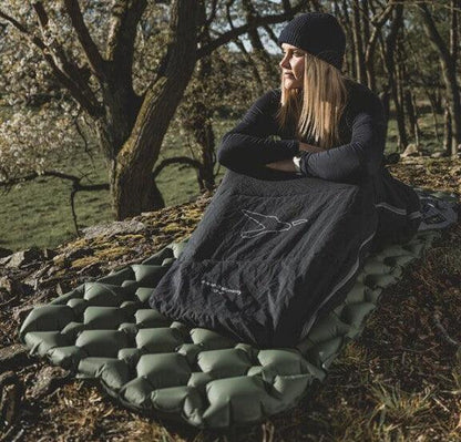 Woman relaxing on green camping mattress in forest, wrapped in black sleeping bag, ideal for outdoor adventures.