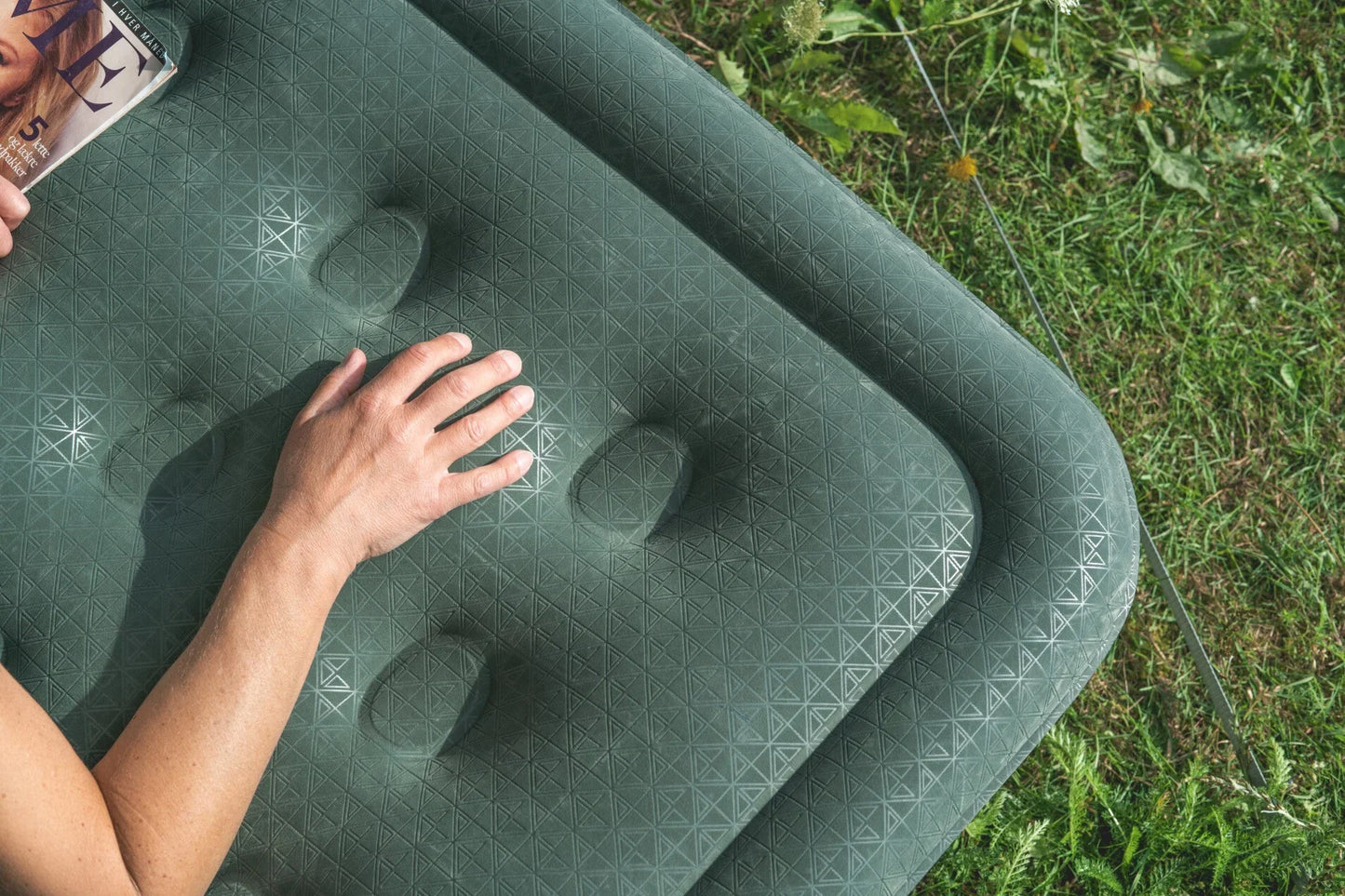 Green inflatable camping mattress with geometric pattern, hand resting on surface, grassy outdoor setting