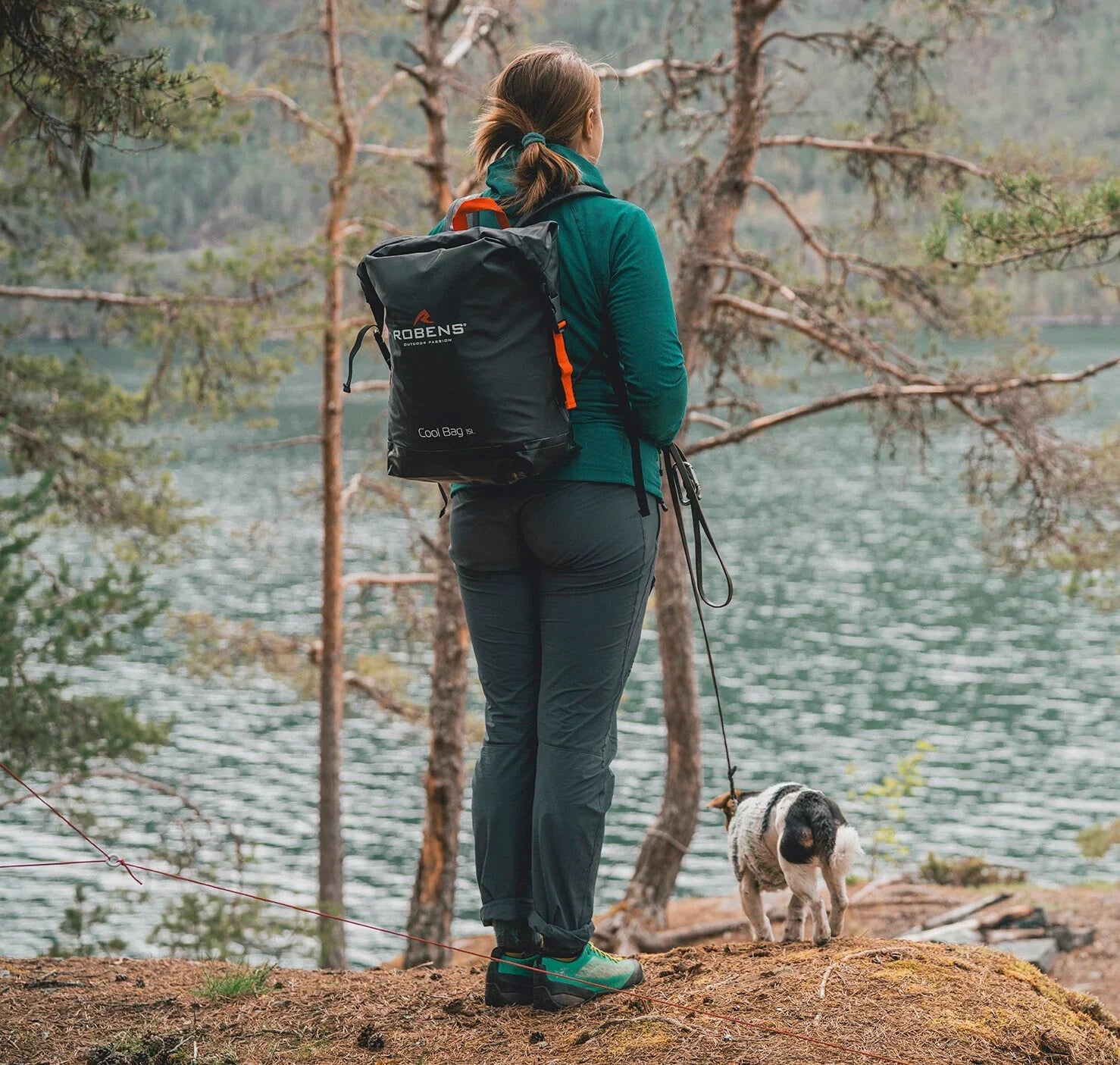Woman hiking with dog near lake, wearing Robens backpack, outdoor adventure gear