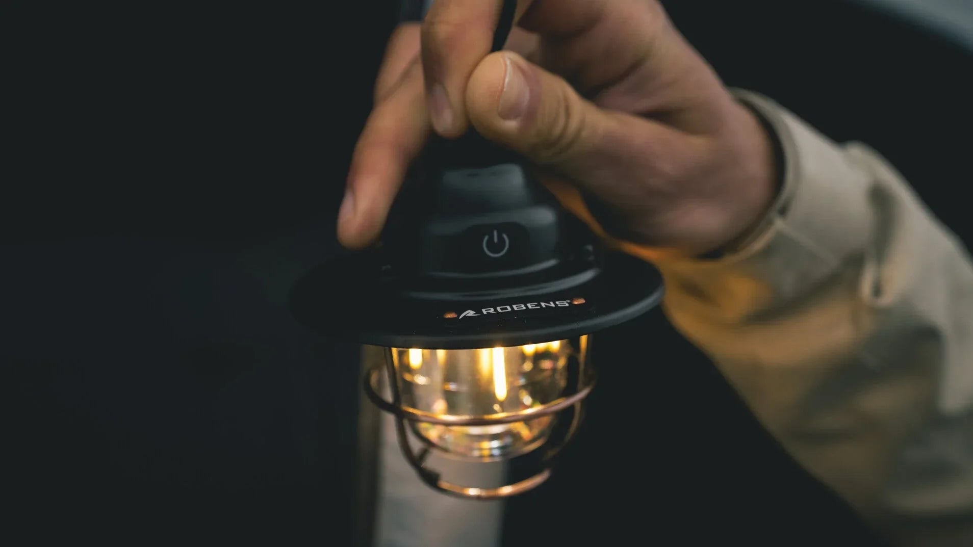 Hand holding lit Robens camping lantern with power button, ideal for outdoor adventures and camping trips.