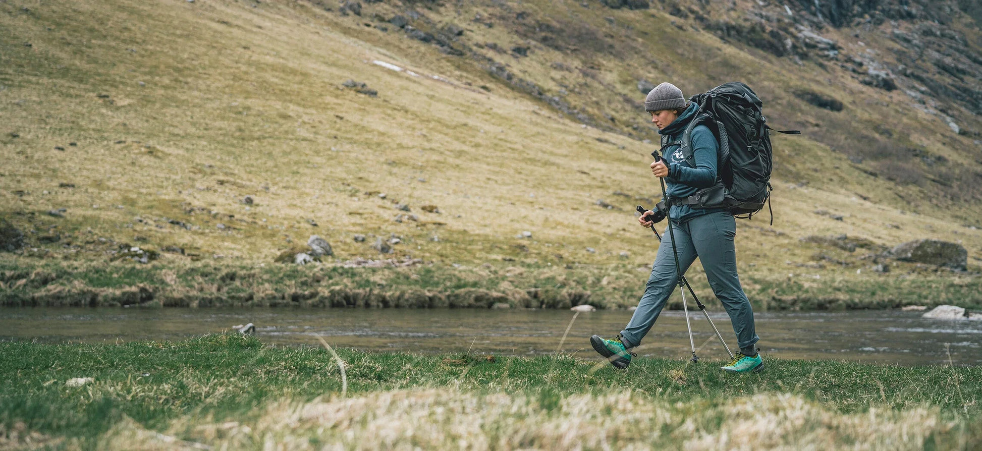 Hiker with backpack and trekking poles walking near river in mountainous terrain, outdoor adventure gear