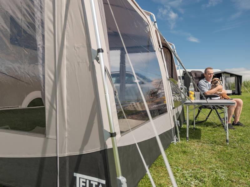 Man relaxing with dog beside caravan awning on grassy field under blue sky