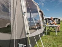 Man relaxing with dog beside caravan awning on grassy field under blue sky