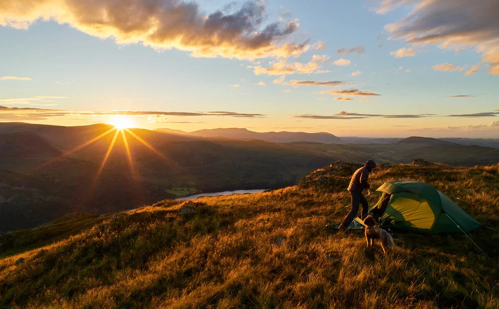Camping at sunset with green tent on hilltop, dog nearby, scenic mountain view