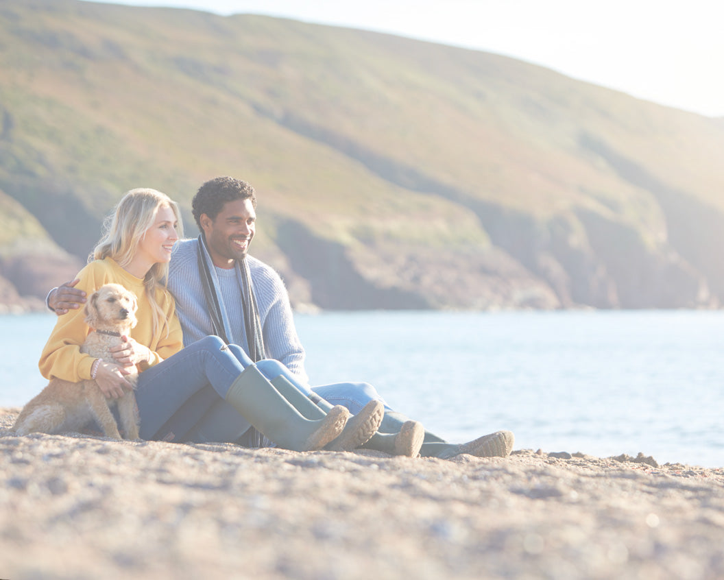A smiling couple sits on a sandy beach with a small dog, with a grassy hill and water in the background. The woman wears a yellow top and blue jeans, and the man wears a light blue sweater and gray scarf.
