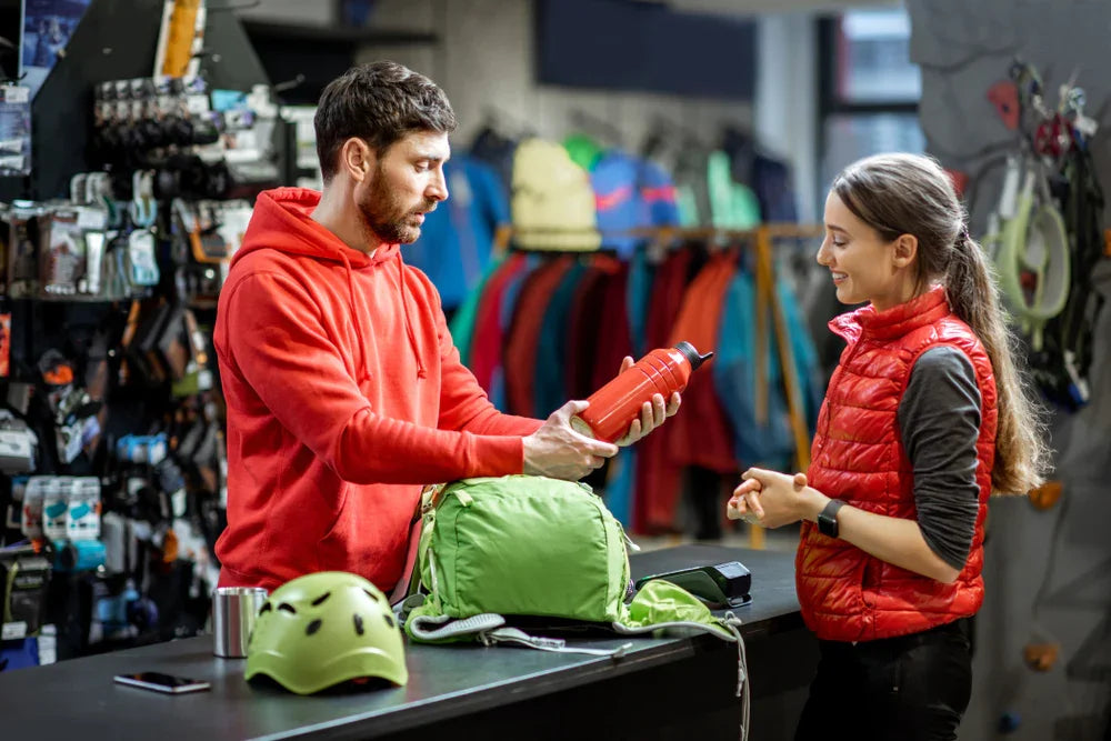 Man holding red water bottle with woman at outdoor gear store, green backpack and helmet on counter