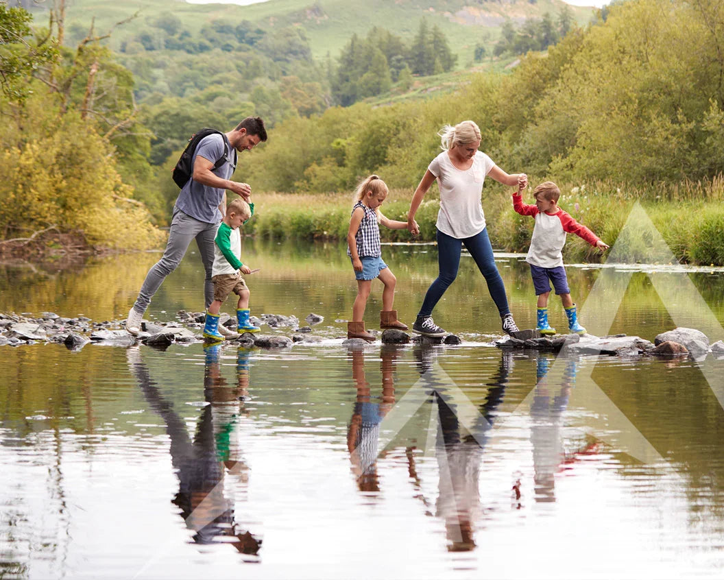 A family of five walks across a rocky stream in a natural outdoor setting with green hills and trees in the background. The adults and children are holding hands and looking down at the rocks they are stepping on.