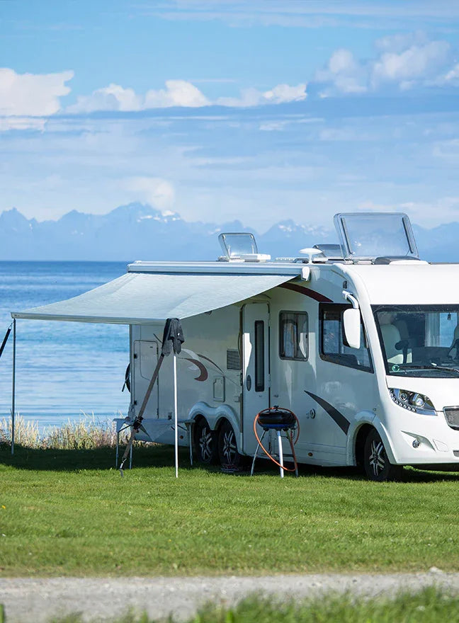 A white motorhome parked on grass by a lake with mountains in the background. The RV has an extended light gray awning, and camping gear is set up beside it under a blue sky with clouds.
