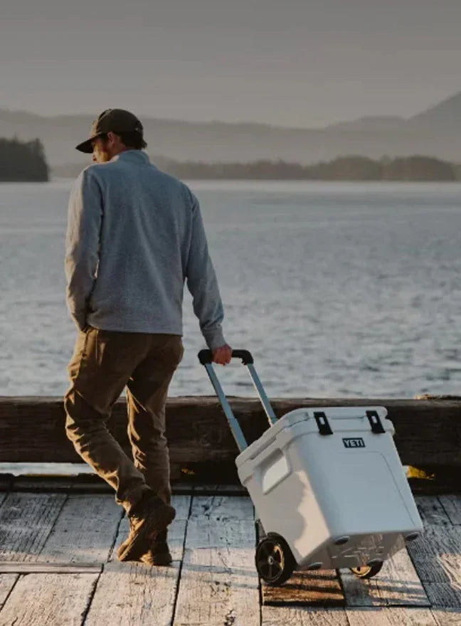 A man seen from behind walks on a wooden dock by the water while pulling a white YETCO brand rolling cooler with an extended handle.