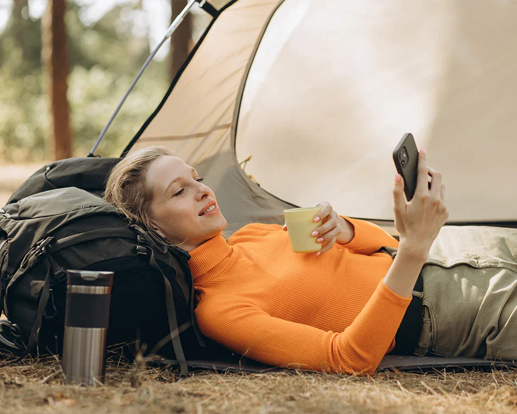 A woman lying on the ground in front of a camping tent, wearing an orange long-sleeve top and khaki pants, holding a smartphone and a mug. A black hiking backpack and a stainless steel tumbler are beside her in a forest setting.