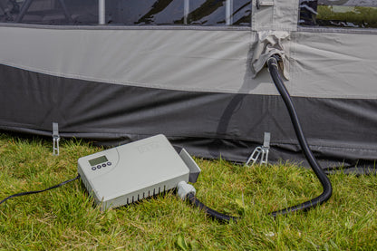 A gray electronic pump controller connected to a black hose, set on green grass next to a gray and black inflatable tent.