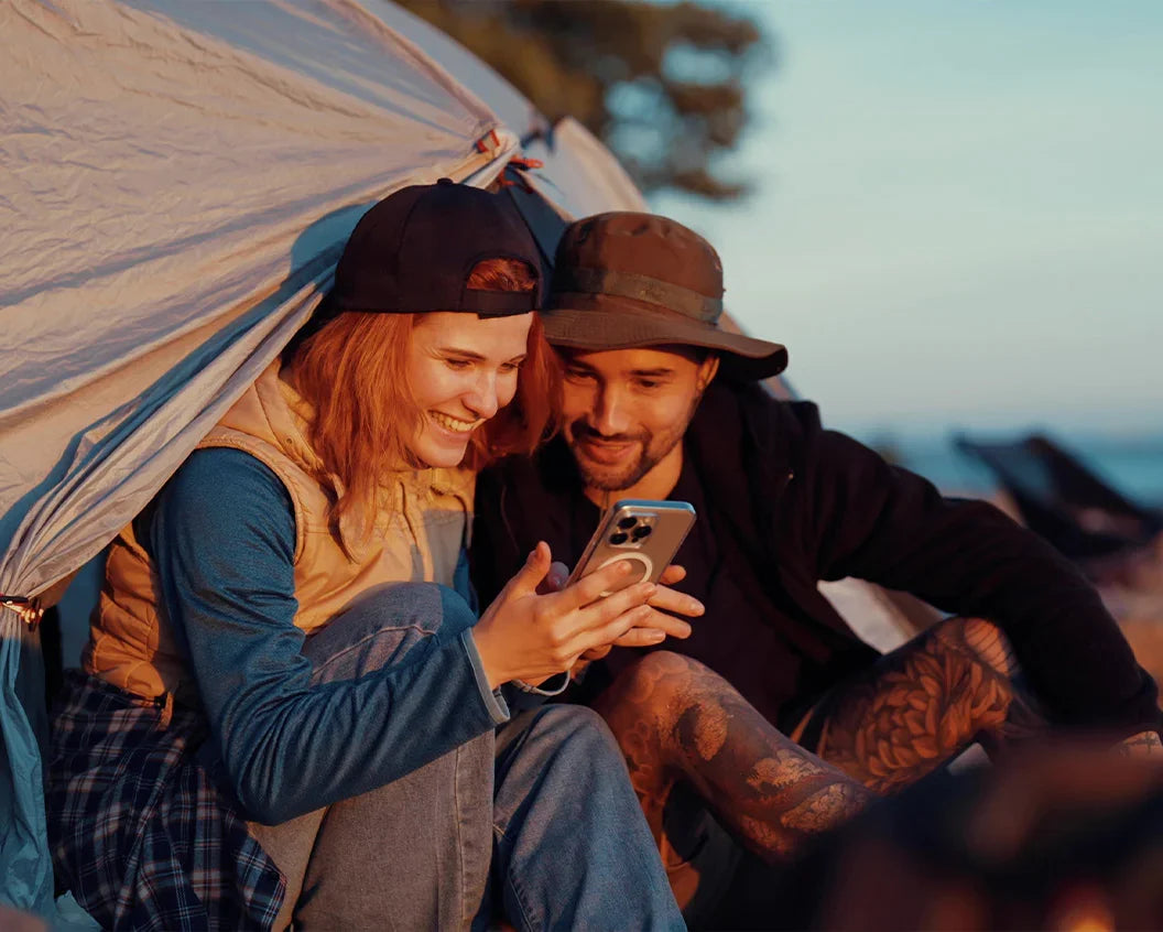 A couple sitting under a white tent at dusk, smiling while looking at a smartphone together. The woman wears a beige vest over a blue long-sleeve shirt and a dark cap, and the man wears a brown wide-brim hat and a dark jacket.