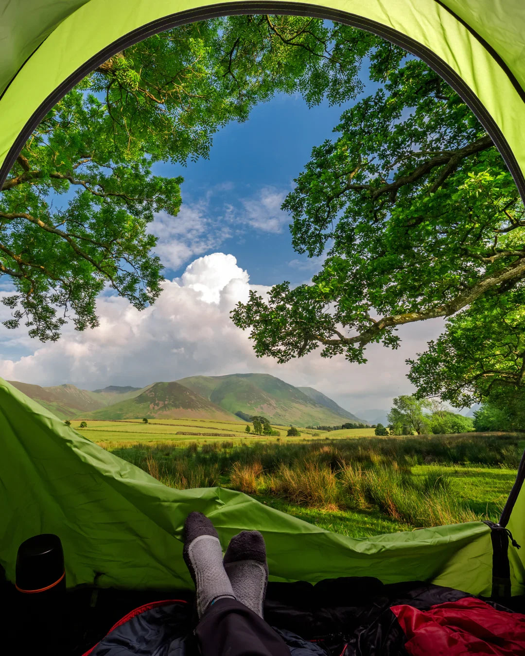 View from inside a bright green camping tent looking out at a scenic landscape with green fields, distant hills, and a blue sky with clouds, with a person's legs visible at the bottom of the tent.