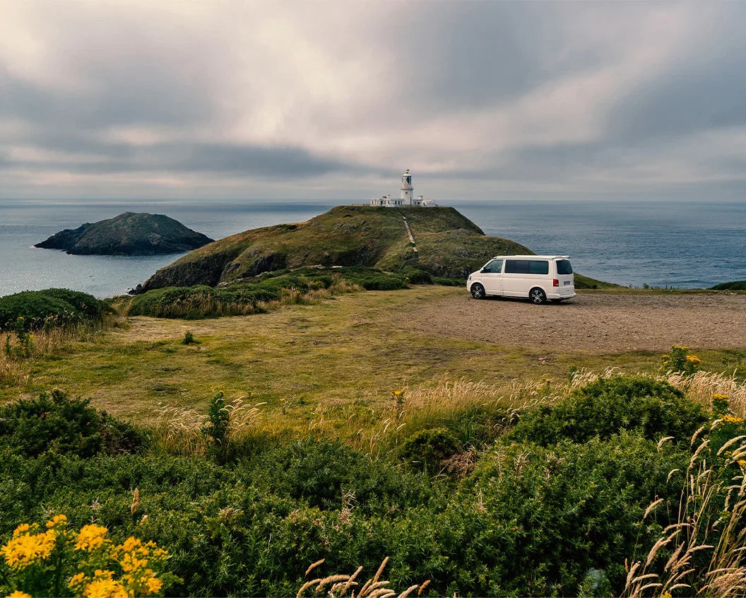 A white minivan parked on a dirt clearing beside a grassy coastal hill with a lighthouse, overlooking the ocean under a cloudy sky.
