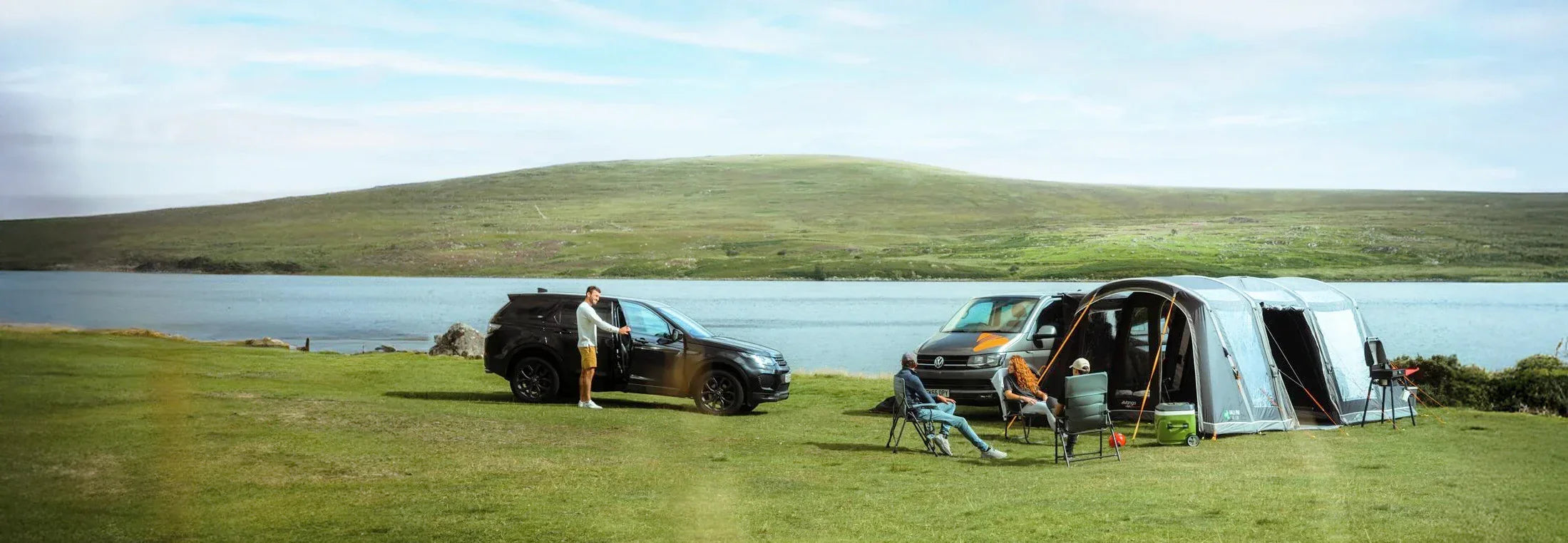 Two vehicles with camping setups by a lake on a grassy area