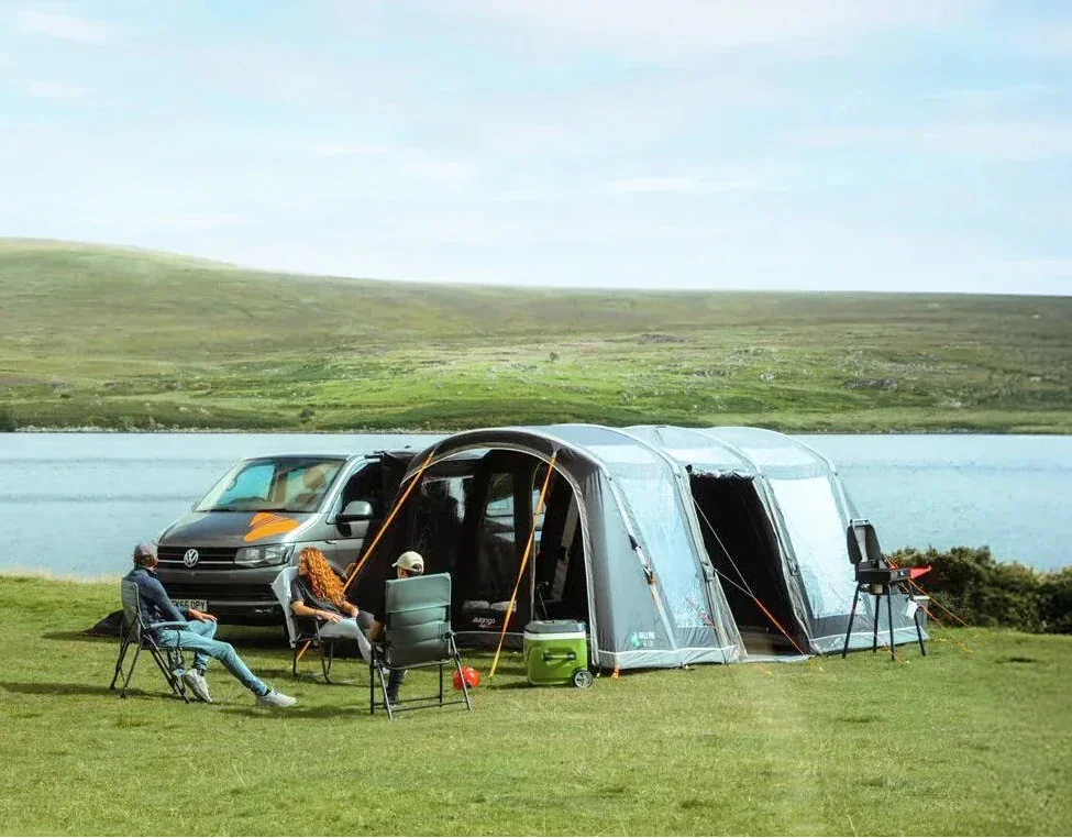 A large silver and white car awning tent is pitched on a grassy lakeside lawn next to a dark gray Volkswagen van, with campers and people relaxing in folding chairs in the foreground.