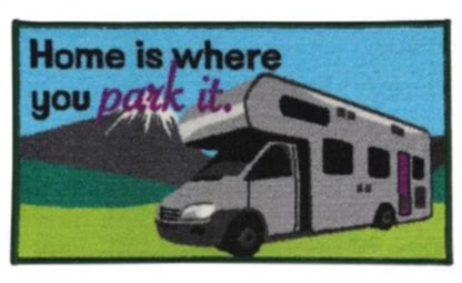 A rectangular woven rug or mat with a colorful outdoor scene showing an RV parked in front of mountains and trees, with the text 'Home is where you park it.' across the top.