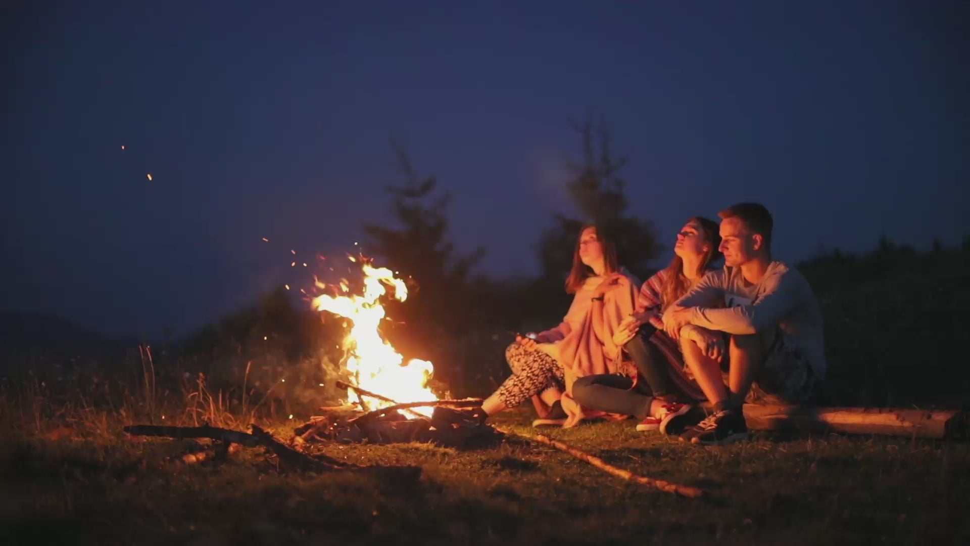 Load video: video showing 3 young people sat outside by a camp fire looking at the night sky