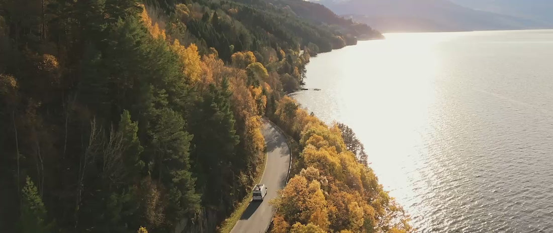 Load video: video showing a motorhome driving along a lake with greenery andtrees on the other side